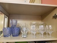 Set of 6 short blue dimpled glasses, 4 margarita glasses, 6 crystal brandy snifters, and short martini glasses displayed inside a cabinet shelf.