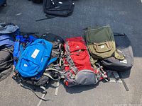 Seven backpacks laid out on the pavement showing various colors and styles including blue, red, black, olive green and navy blue.