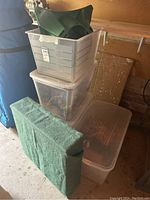 Three plastic storage bins stacked, one with a green plastic Christmas tree stand on top and a foldable green fabric-covered display platform leaning in front