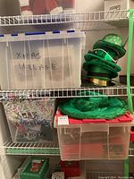 Plastic totes labeled 'Xmas Village' and 'Christmas Stockings & Hangers' on basement shelves alongside numerous green St. Patrick's Day hats stacked in various styles.