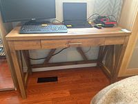 Desk with laminate veneer top and wood grain finish, showing central drawer and cross-braced legs. Items on desk include keyboard, laptop, monitor, and power tools but are not included.