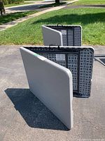 Two 6-foot gray plastic folding tables folded in half standing upright on blacktop, showing front side and back side construction.