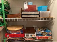 Shelf with multiple board games stacked including Mastermind, Scattergories, Scrabble, Survivor and Ganze bord, plus visible wooden game boards on top shelf