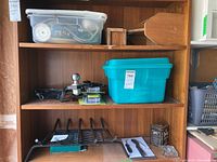 Three shelves holding assorted hardware including a clear plastic bin, turquoise bin, black metal fireplace grate, small hand tools, and a trailer hitch ball mount.