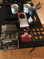 Overview of lot items including wallet, watch in tin case, enamel mugs, manicure set, collectible coins, and sunglasses on a table.