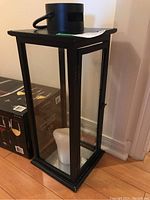 Front and side view of rectangular black metal lantern with clear glass panels and white candle inside, placed on wooden floor next to a wall.