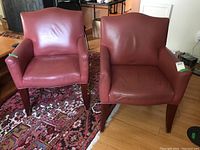 Front view of two matching red leather armchairs on a decorative rug, showing armrests, seat cushions, and wooden legs.