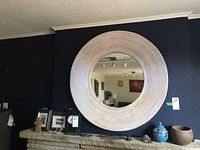 Round wood frame wall mirror hanging on a dark navy blue wall above a mantle with various decorative items.