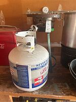 Propane tank with attached burner on wooden shelf, showing the propane tank's label and handle.