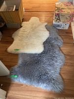 Two sheepskin rugs shown overlapping on a wooden floor, one off-white and the other gray, with green tags attached.