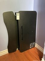 Black standing desk riser folded and leaning against wall, showing top and bottom surface.