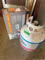 White 20 lb Flame King propane tank next to Remington tank top heater box on concrete floor.
