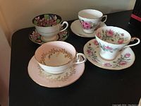 Four tea cups and saucers showing different floral designs and gold trim on porcelain, photographed on black surface.