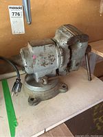 Metal bench vise positioned on a white table showing side profile, the handle, and power cord.