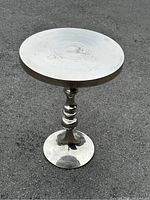 Round aluminum accent table on outdoor surface, showing wear patterns on top and polished finish on pedestal base
