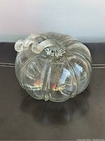 Clear glass pumpkin with curled detailed stem, shown from the front on a dark surface.