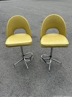 Front view of two yellow vinyl vintage bar stools with chrome bases on outdoor pavement showing overall condition and design.