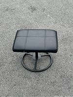 Black leather padded ottoman foot rest stool with visible stitched square pattern on top, round metal footed base shown from top-down angle on asphalt.