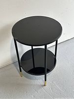 Front angled view of round black side table with two shelves and slender black legs ending with gold caps