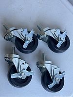 Photo showing 4 caster wheels with metal mounting bolts and brake handles angled upwards on a white surface.