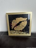 Front view of golden lips wall art with gold sequin decoration inside a metallic gold frame on a black background.