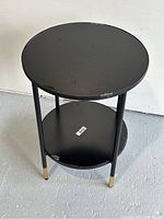 Front-side view of black IKEA round glass side table showing glass top, lower shelf and metal legs with brass feet caps.
