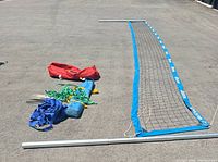 Full volleyball net laid out with blue borders and white ropes, poles attached. Setup equipment spread out on asphalt.