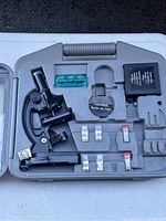 Full view of microscope set in gray plastic case showing black microscope, petri dish, slide box, prepared slides, and small containers.