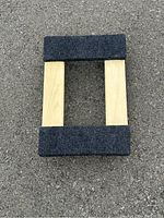 Furniture mover's dolly viewed from above showing wooden frame with gray carpet padding on the surface and edges, placed on pavement.