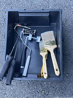 Top view photo of 3 paint rollers, 3 paint brushes in a black rectangular plastic tray on an asphalt surface.