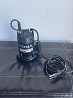 Front view of black Rona submersible pump with attached black electrical cord and plug coiled beside it on dark surface.