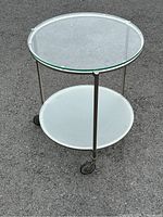 Front view of the stainless steel table showing glass top and lower glass shelf, mounted on three vertical legs with wheels.