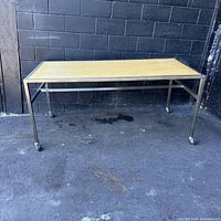 Front view of a rectangular mobile work table with wood top and metal frame against a black brick wall, showing caster wheels and lower supports.
