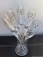 Group of six clear glass toasting flutes decorated with gold stars arranged in a clear glass holder, shown from an angle.