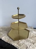 Front angle view of the golden three-tiered serving tray on a neutral textured surface.