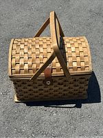 Woven picnic basket with domed lid and dual wooden handles, natural light wood color, taken on pavement outdoors.