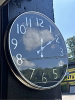 Round wall clock mounted outdoors with reflective glass face showing sky and clouds, chrome or metallic rim, black face, and white numerals.