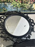 Black ornate oval wall mirror with intricate plastic frame, shown hanging on wall.