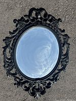 Front view of oval mirror with ornate black plastic frame showing detailed scrollwork and floral design.