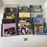 Photo showing 15 music CDs from various artists, including The Hollies, Neil Diamond, Loverboy, Ritchie Valens, Bill Haley, Steve Winwood, and others arranged on a shelf.