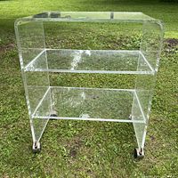 Front view of the clear lucite cart with three shelves and four wheels on grass background.