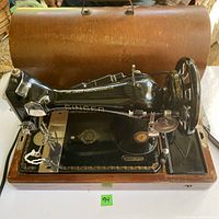 Front view of the Singer Centennial Sewing Machine on its wooden base and carrying case, showing black and gold decorative machine with power cord and pedal.