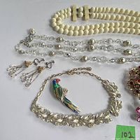 Photo shows an assortment of vintage costume jewelry including a faux pearl multi-strand bracelet, rhinestone floral bracelet, clear crystal bead necklace, colorful parrot brooch, and clip-on earrings.