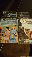 Four original Nancy Drew books laid out on a wooden surface showing front covers of each book with title and author Carolyn Keene visible.