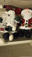 Close-up of two Santa figurines, one traditional red and white, the other textured white with curly beard and pom-pom accents