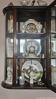 Front view of curio cabinet section displaying Precious Moments figurines, decorative plates, small crystal figurines, and tabletop clock.