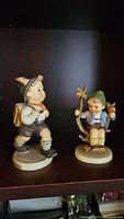 Both Hummel figurines, Apple Tree Boy and School Boy, displayed on a shelf, showing full view of each figurine.