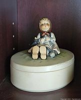 Photo showing the full round cream-colored porcelain trinket box with the Hummel figurine lid featuring the seated girl, cat and bird.