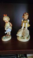 Both Hummel figurines, Little Gardener and Goose Girl, displayed side by side on a wooden surface with dark background.
