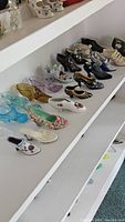 Overview of the assortment of 14 miniature shoe collectibles displayed on white shelves including glass, porcelain, and ceramic materials in varying colors and designs.